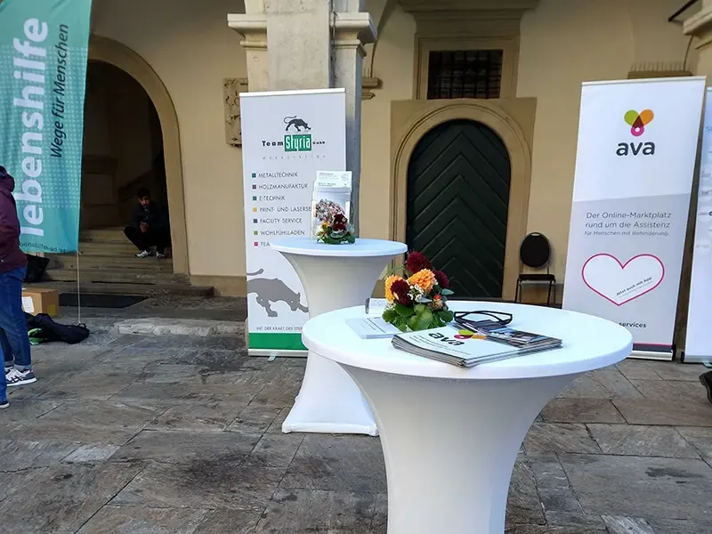 Zwei Stehtische mit Blumen und Informationsmaterial vor Bannern von Lebenshilfe und ava beim Sozialtag im Landhaushof Steiermark