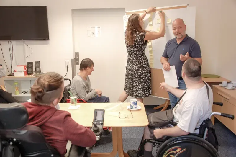 Fünf Personen, darunter drei im Rollstuhl, arbeiten gemeinsam an einem Whiteboard mit Haftnotizen in einem Besprechungsraum.