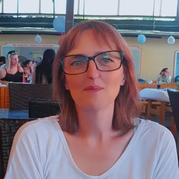 Frau namens Manuela mit roten Haaren und schwarzer Brille, sitzt in einem Restaurant mit anderen Gästen im Hintergrund