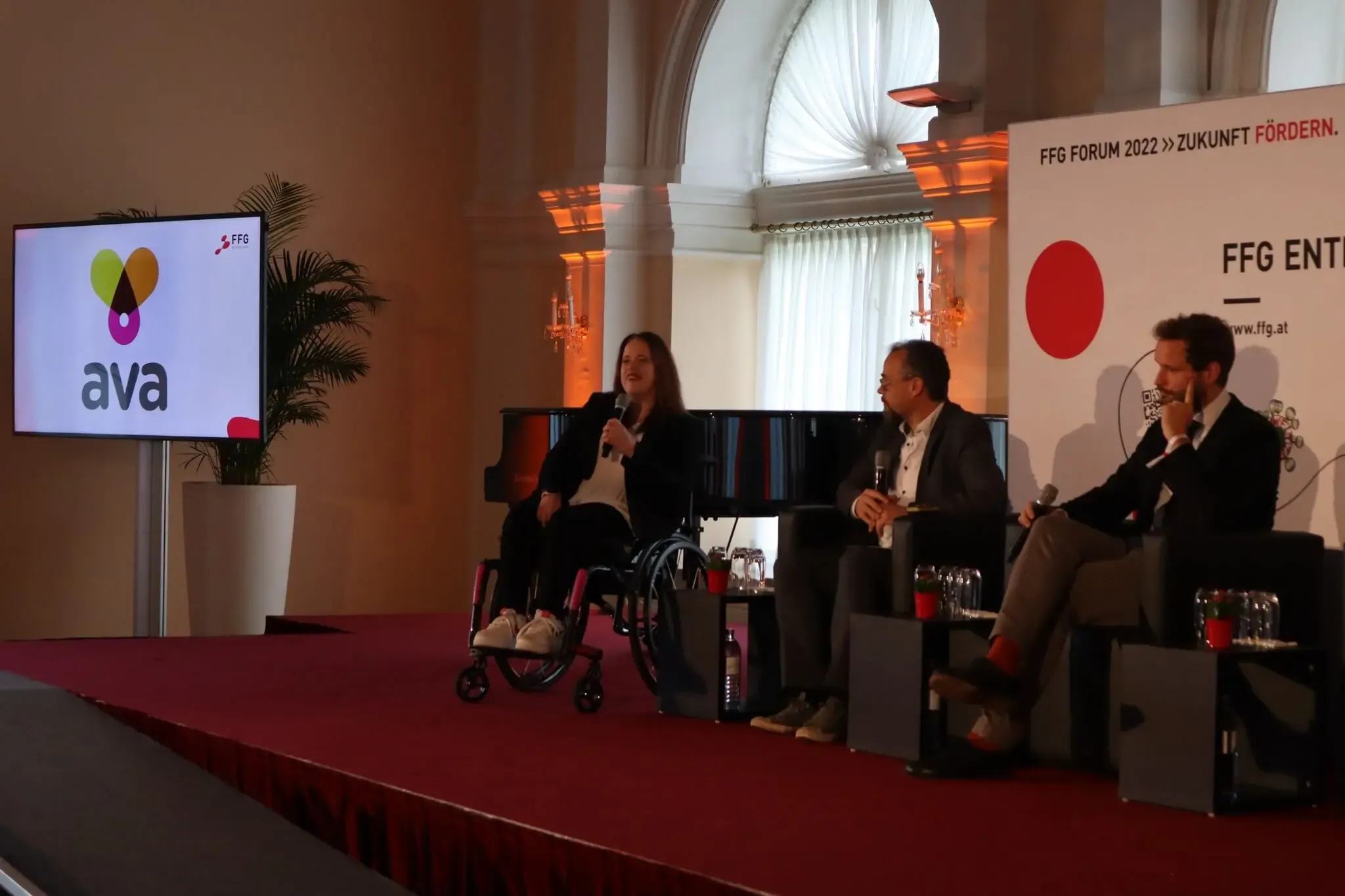Melanie Wimmer im Rollstuhl spricht auf der Bühne des FFG Entrepreneurship Day 2022, begleitet von zwei weiteren Personen bei einer Podiumsdiskussion