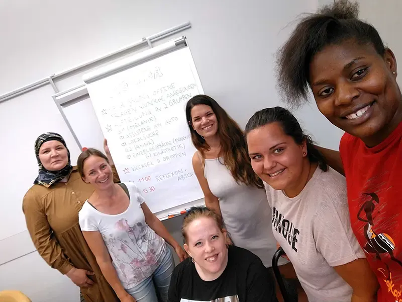 Sechs Frauen unterschiedlichen Alters und Herkunft lächeln in einem Klassenraum vor einem Flipchart mit handgeschriebenen Notizen