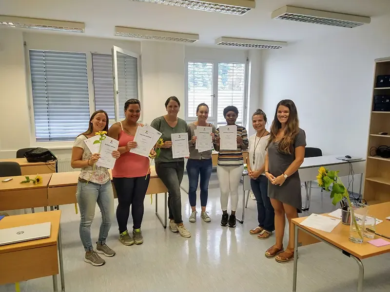 Anna Grill (ganz rechts) mit fünf Ausbildungsteilnehmerinnen, die Zertifikate und Sonnenblumen in einem Klassenraum halten