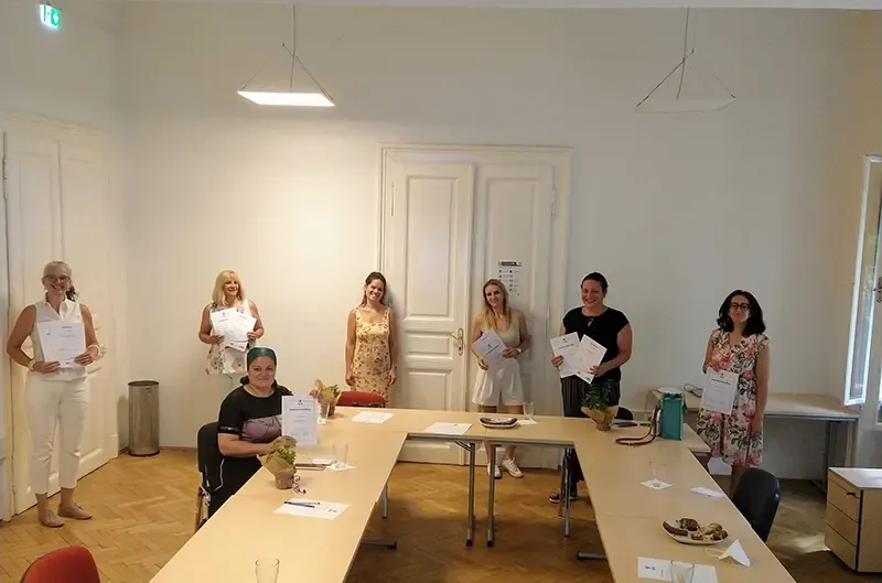 Anna Grill im geblümten Kleid stehend mittig mit sechs Ausbildungsteilnehmerinnen in einem hellen Raum, alle halten Zertifikate